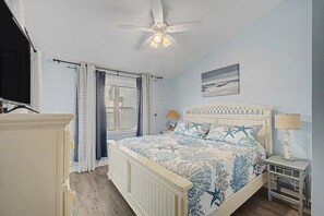 Ein Schlafzimmer mit Bett, Deckenventilator, Fenster mit Vorhängen, Nachttisch mit Lampe und einem Bild an der Wand.