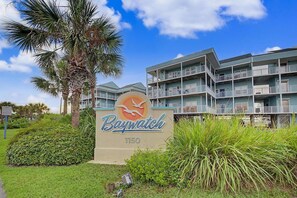 A hotel sign with a bird and sun logo, surrounded by greenery and palm trees, with a multi-story building in the background. - Escape by the bay! Baywatch E10 (Pensacola Beach)