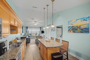 A kitchen with wooden cabinets, a granite countertop, and a dining area with wicker chairs. - Escape by the bay! Baywatch E10 (Pensacola Beach)