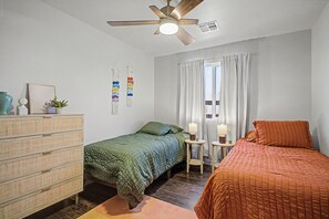 Ein Schlafzimmer mit zwei Betten, einem Deckenventilator, einem Fenster mit Vorhängen und einer Kommode mit Schubladen.