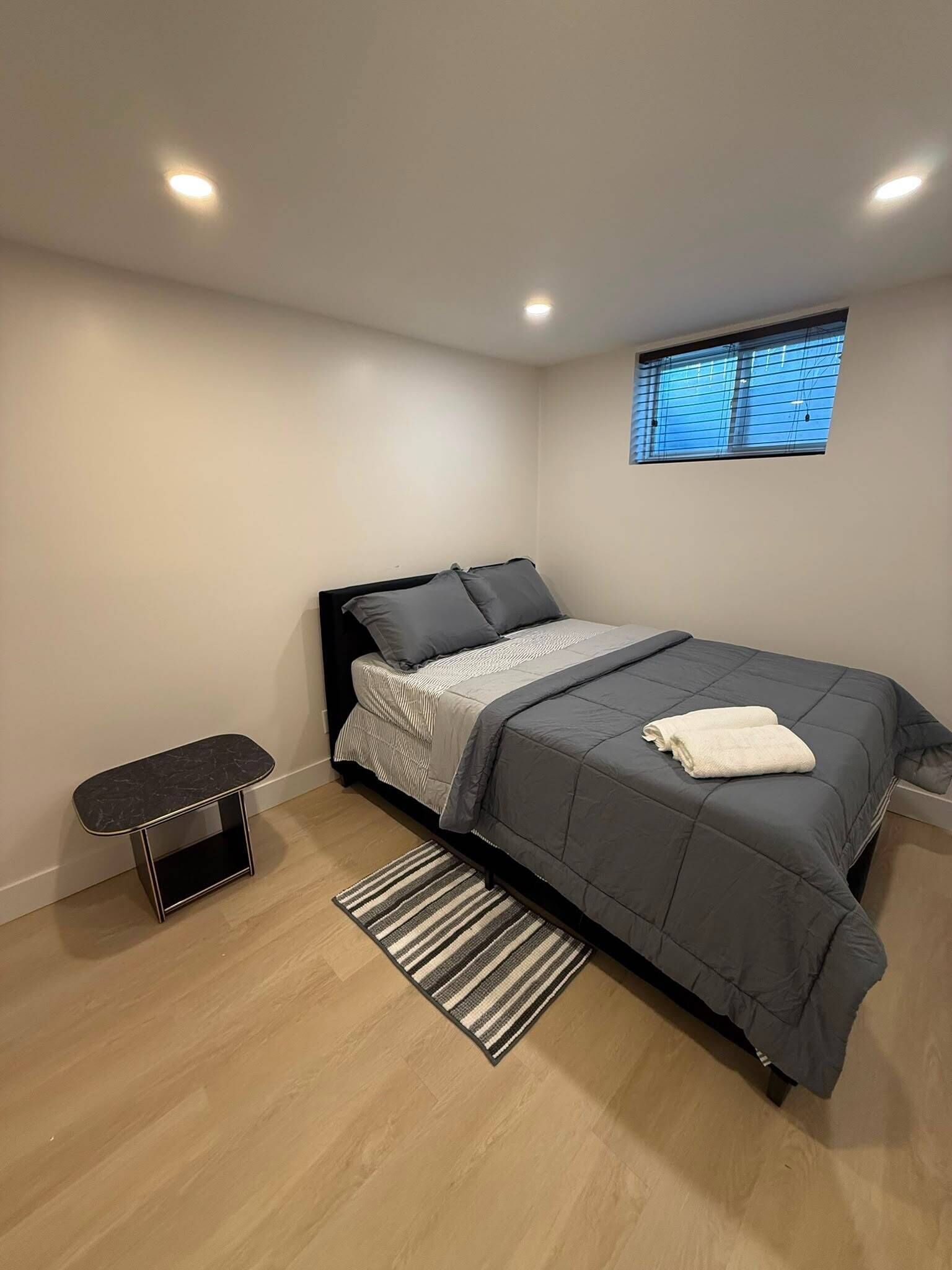 A bedroom with a bed, a bedside table, a window, and a small bench.
