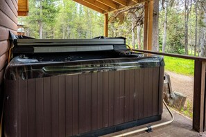 A hot tub in a covered outdoor area with a wooden deck and a view of a forest. - Cozy Island Park Cabin, Hot Tub, Snowmobile Trails (Island Park)