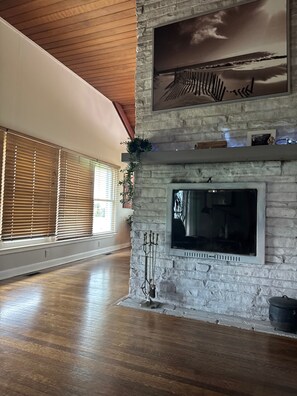 A spacious room with a stone fireplace, wooden flooring, and large windows with blinds. - Peaceful Four-Bedroom Country Retreat with Hot Tub & InfraredSauna (Parkville)