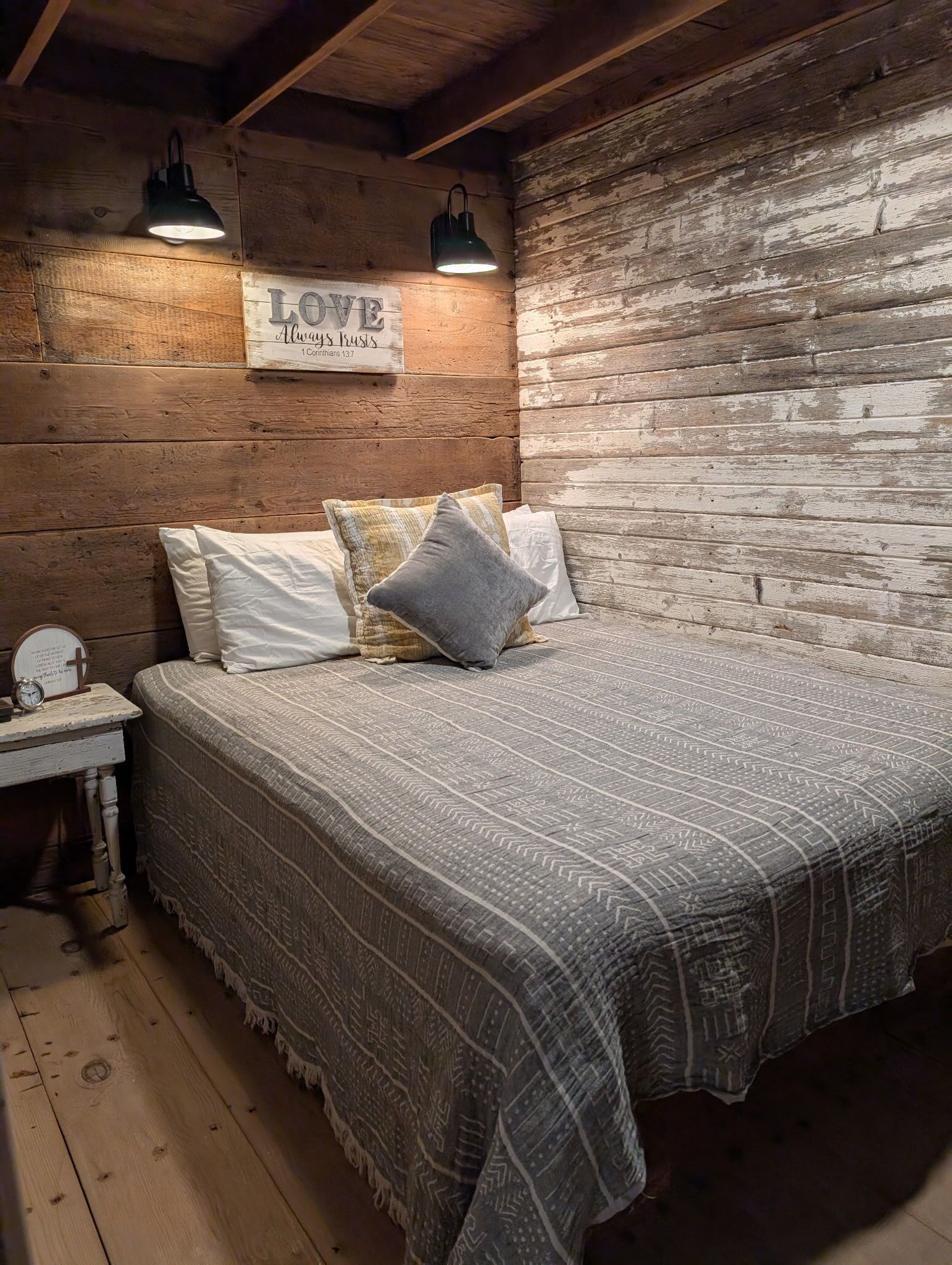 A rustic bedroom with a wooden wall, a bed with patterned bedding, and a wall-mounted sign.