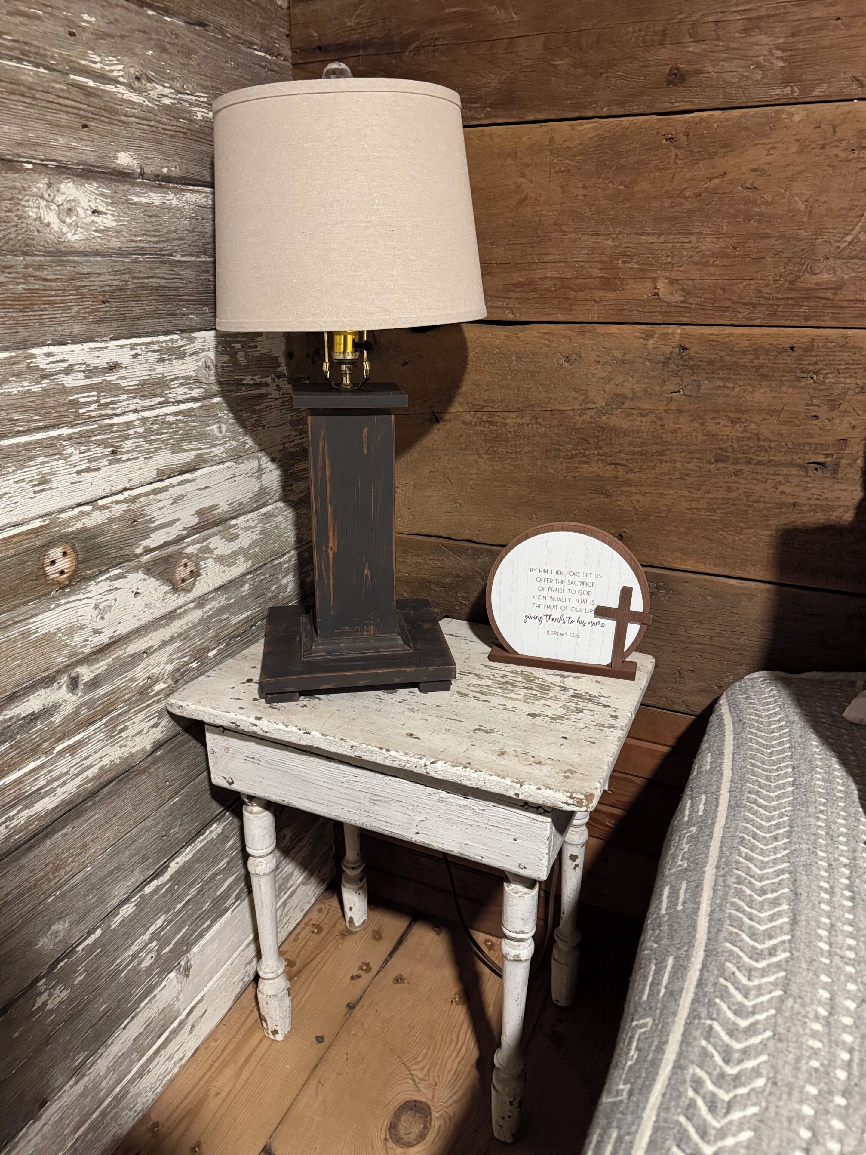A rustic wooden wall with a small table featuring a lamp and a religious plaque.