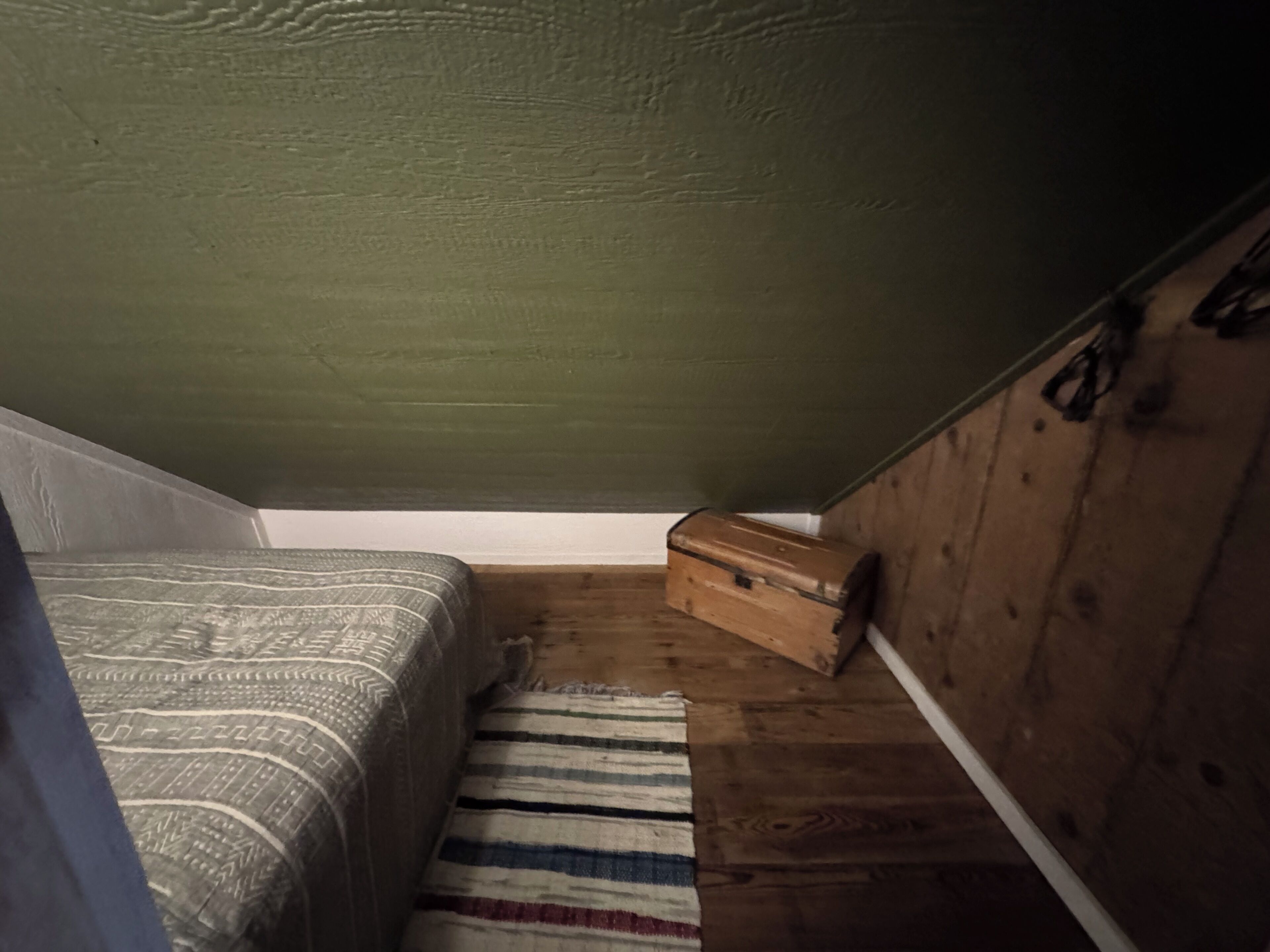 Un pequeño ático tenuemente iluminado, con techo inclinado, una cama individual, un cofre de madera y un tapete a rayas.