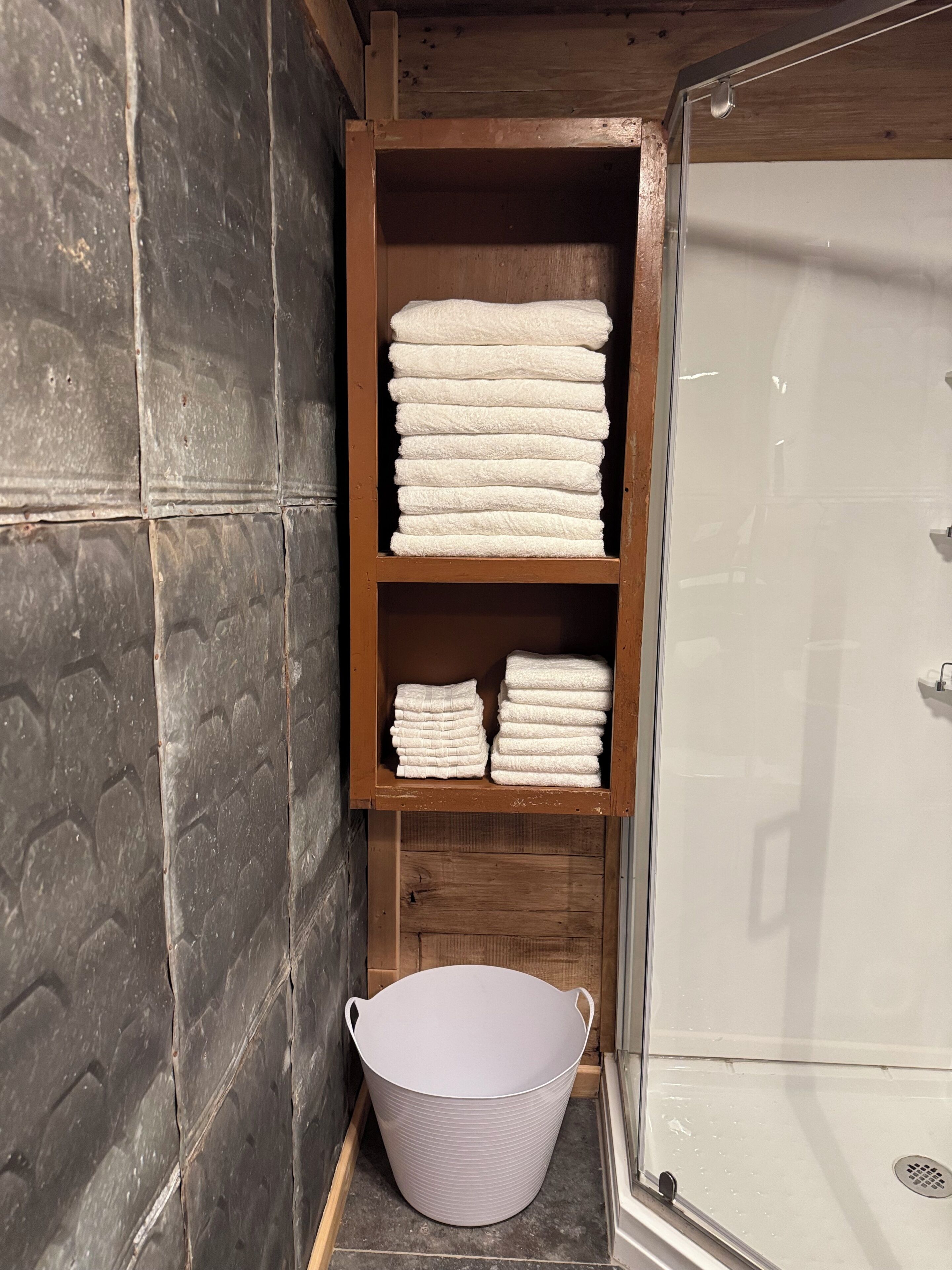 Casa de banho com prateleira de madeira com toalhas dobradas, um balde branco e zona de duche.
