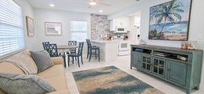 A living room with a sofa, a coffee table, and a dining area with chairs and a table. There is a kitchen with white cabinets and a microwave. A painting of a beach scene hangs on the wall. - Private Waterfront House • Private Dock • 2 Min Walk to Beach (Indian Shores)