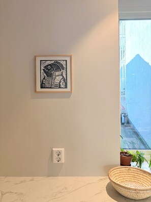 Une œuvre d’art encadrée accrochée au mur, une prise de courant, une plante en pot et un panier tressé posés sur un comptoir en marbre.