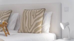 Una cama bien hecha con ropa blanca y almohadas con estampado de cebra, una mesita de noche con una lámpara, y un taburete de madera con un vaso y una botella.