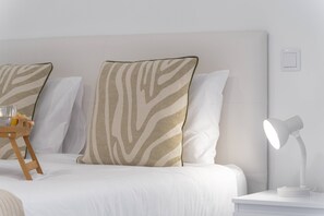 A neatly made bed with white bedding and zebra-patterned pillows, a bedside table with a lamp, and a small wooden stool with a glass and a bottle. - Branco's Apartment 03 by GALMI (Ponta do Sol)