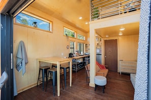 Un intérieur chaleureux avec une table à manger en bois, des tabourets de bar, un coin cuisine et un petit espace salon comprenant une chaise et un pouf.