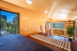 Une chambre dotée d’un grand lit, d’un plancher en bois, d’une fenêtre donnant sur la verdure et d’un fauteuil agrémenté d’un coussin au design original.