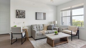 A modern living room with a grey sofa, a coffee table, and two armchairs. There is a painting on the wall and a window with a view. - Landing | Modern 2BD, Gym, Clubhouse (Dallas)