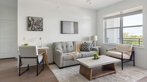 Ein modernes Wohnzimmer mit einem grauen Sofa, einem Couchtisch und zwei Sesseln. An der Wand hängt ein Gemälde und das Fenster bietet einen Ausblick.