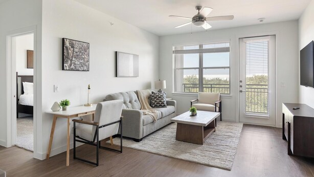 A modern living room with a sofa, coffee table, and a desk with a lamp. There is a ceiling fan, large windows, and a door leading outside. - Landing | Modern 2BD, Gym, Clubhouse (Dallas)