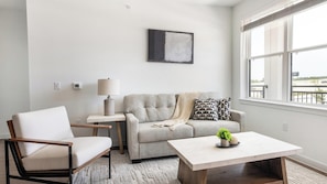 A living room with a sofa, a chair, a coffee table, a lamp, and a painting on the wall. - Landing | Modern 2BD, Pool, Gym (Dallas)