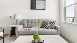 En stue med en grå sofa, et lille sofabord med en lampe, et sofabord med dekorative genstande, og et vindue med udsigt.