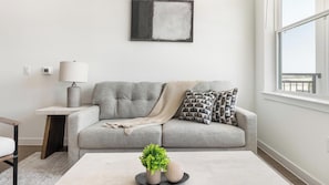 A living room with a grey sofa, a small side table with a lamp, a coffee table with decorative items, and a window with a view. - Landing | Incredible 2BD, Courtyard, Clubhouse (Dallas)
