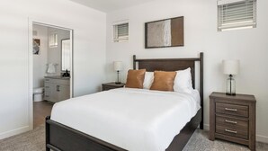 Et hotelværelse med en stor seng, et natbord med lampe, et kommode, et badeværelse med toilet og håndvask, samt et maleri på væggen.