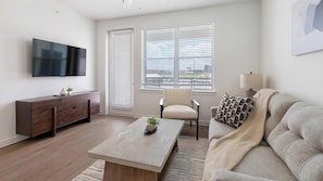 A modern living room with a flat-screen TV, a wooden coffee table, a beige sofa, and a window with blinds. - Landing | Amazing 1BD, Clubhouse, Pool (Dallas)