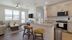 A modern kitchen with a central island, a dining area with a sofa and coffee table, and a living room with a TV. - Landing | Amazing 1BD, Pool, Courtyard (Dallas)