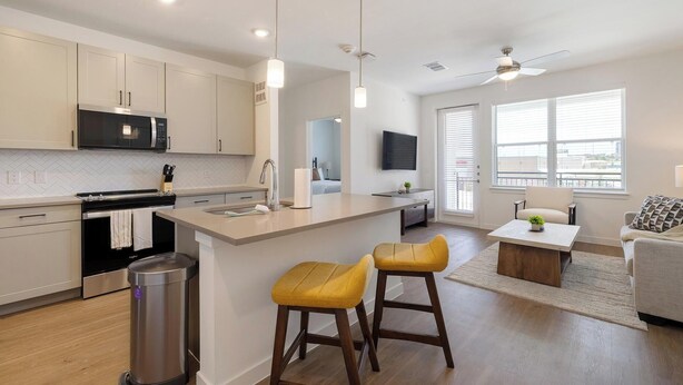 Una cocina moderna con una isla central, taburetes de bar, un comedor con mesa y una sala de estar con sofá y televisor.