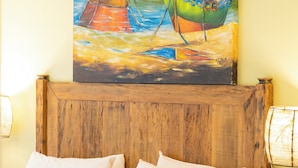 Une chambre d’hôtel avec une tête de lit en bois, un lit recouvert de draps blancs, plusieurs oreillers, une table de chevet avec une lampe et un tableau représentant des bateaux accroché au mur.