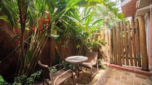 Une terrasse dotée de meubles en osier et d’une table, entourée d’une végétation tropicale luxuriante et d’une clôture en bois.
