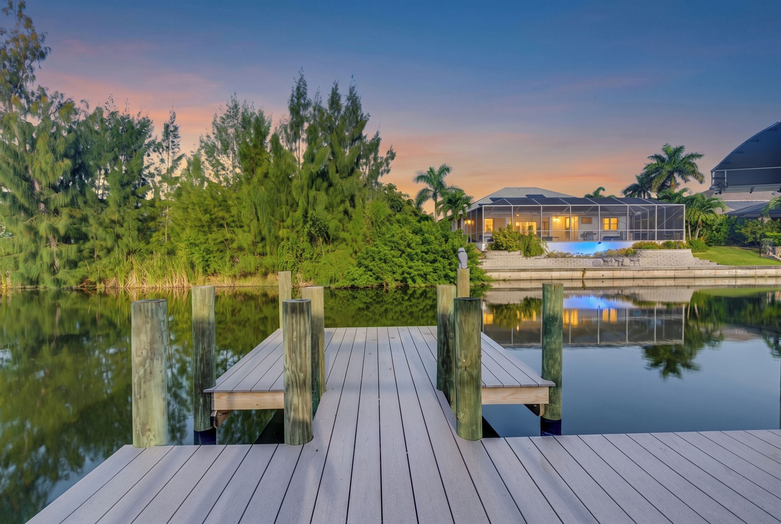 A wooden dock leading to a house with a pool, surrounded by trees and a calm lake.