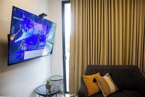 Un televisor de pantalla plana montado en una pared, una mesita redonda con una planta, un sofá con cojines y una ventana con cortinas.