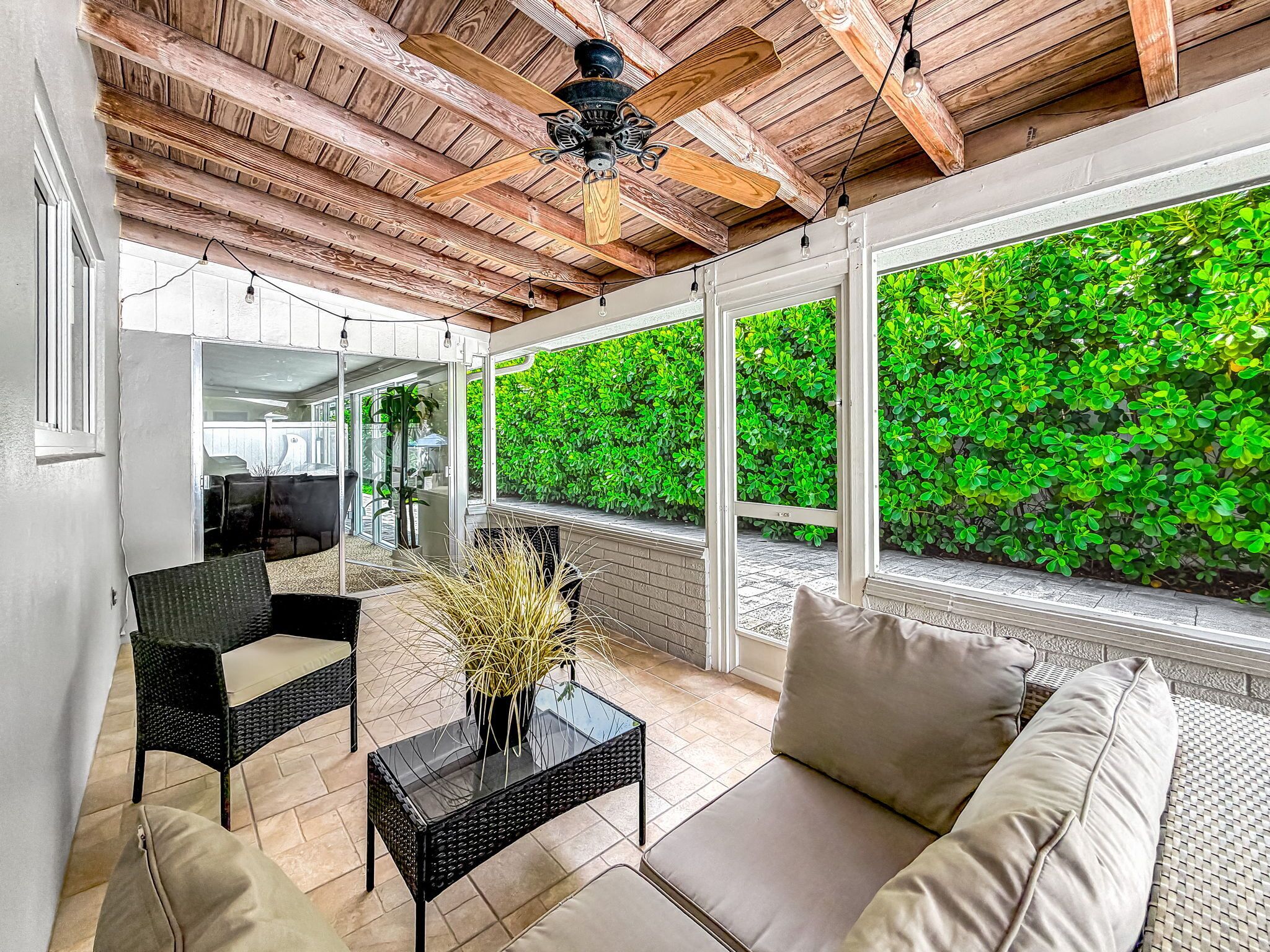 A sunlit patio with a ceiling fan, a sofa, a table, and a view of lush greenery.