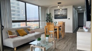 Una sala de estar moderna con sofá, mesa de comedor, sillas y vistas a la ciudad a través de amplios ventanales.