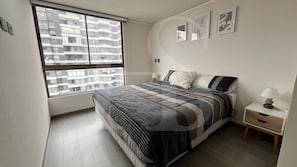 Un dormitorio con cama, mesita de noche, lámpara y ventana con vistas a edificios.