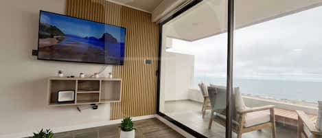 A modern living room with a flat-screen TV, wooden panel wall, and a balcony with a view of the sea.