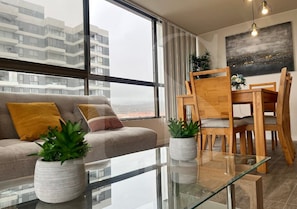 A modern living room with a sofa, a glass coffee table, and a dining area with wooden chairs.