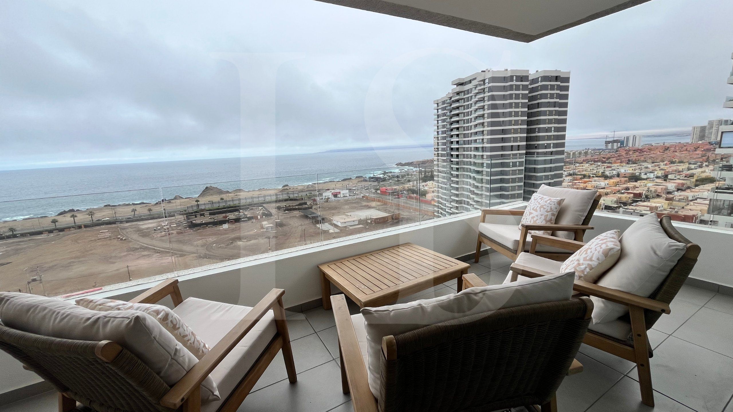 Varanda com vista para a praia e para a paisagem urbana, mobiliada com cadeiras de vime e uma mesa pequena.