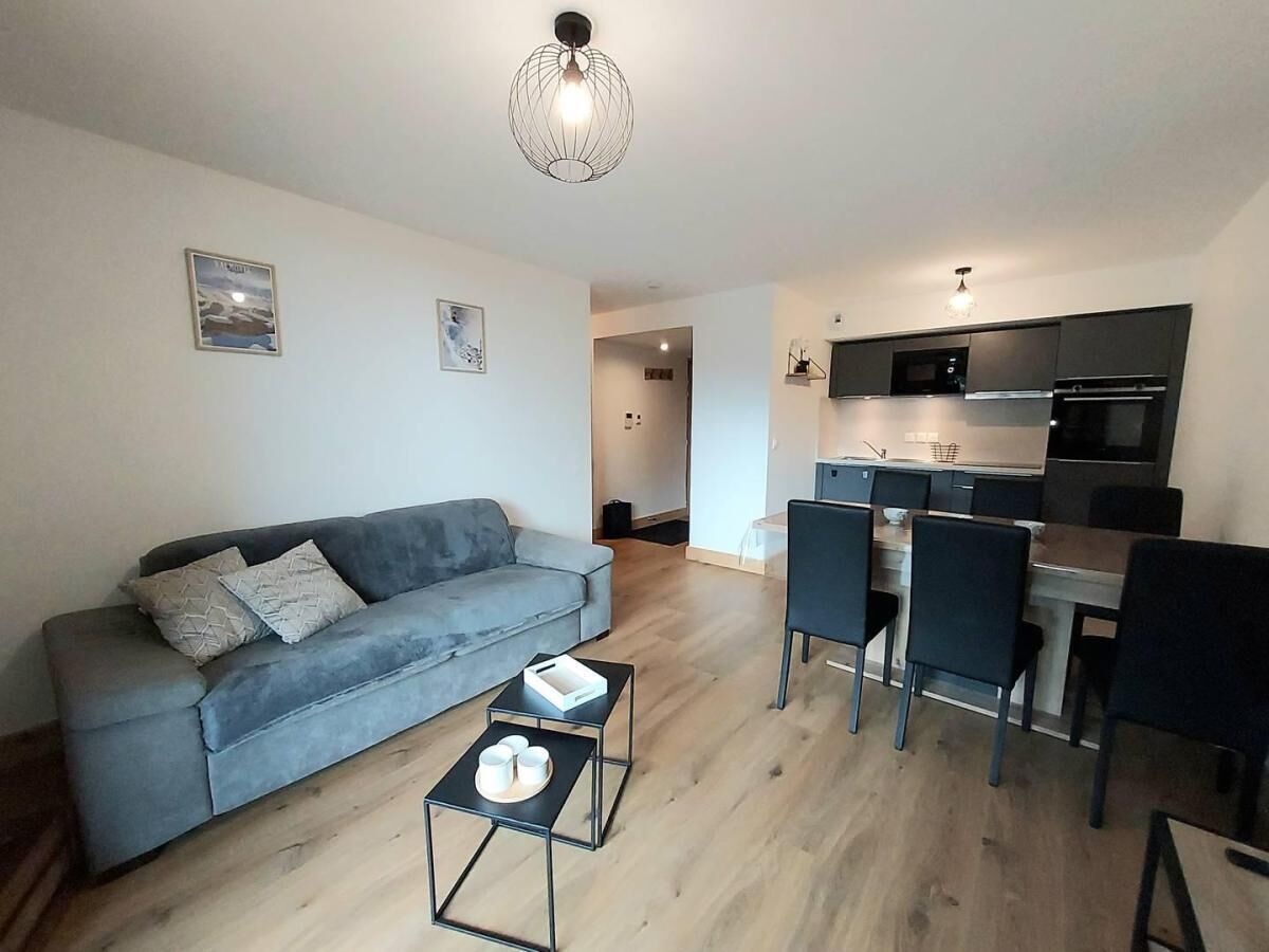 A modern living room with a grey sofa and a small coffee table, a dining area with chairs, and a kitchen with dark cabinets and appliances.