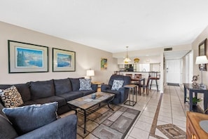 A living room with a sofa, coffee table, and chairs, a dining area with a table and chairs, and a kitchen in the background. - Edgewater Golf Villas (Panama City Beach)