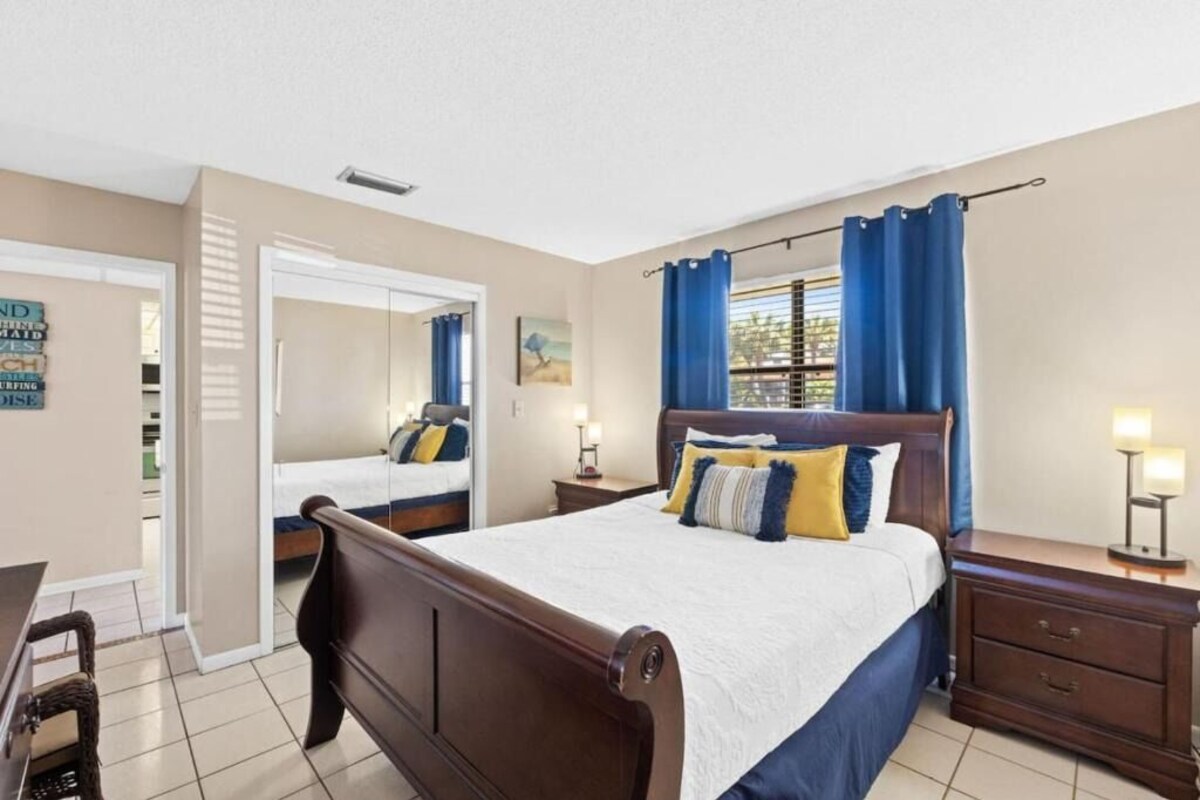 A bedroom with a bed, a nightstand, a dresser, a window with curtains, a mirror, and a door leading to another room.