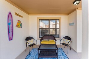 A covered patio area with two wicker chairs, a small table, and a window with blinds. - Horizon South (Panama City Beach)