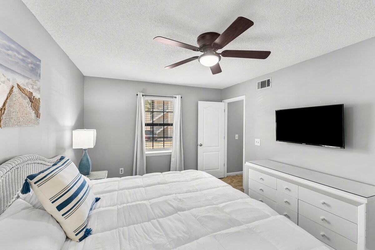 A bedroom with a bed, a ceiling fan, a TV, and a window with blinds.