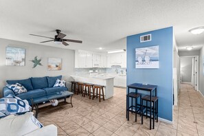 A modern living room with a blue sofa, a glass coffee table, and a kitchen area with white cabinets and a blue accent wall. - Horizon South (Panama City Beach)