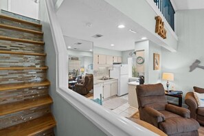 A modern interior with a staircase, a living area with a sofa and armchairs, and a kitchen with white cabinets. - Nautical Watch (Panama City Beach)