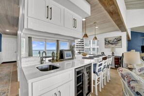 A modern kitchen with white cabinetry, a large island, and a dining area with white chairs. - Nautical Watch (Panama City Beach)