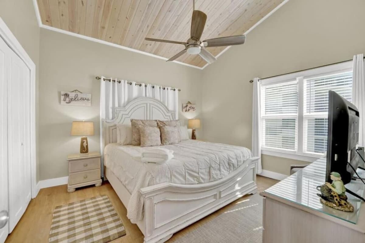 A bedroom with a bed, bedside tables, a ceiling fan, a window with blinds, and a television.