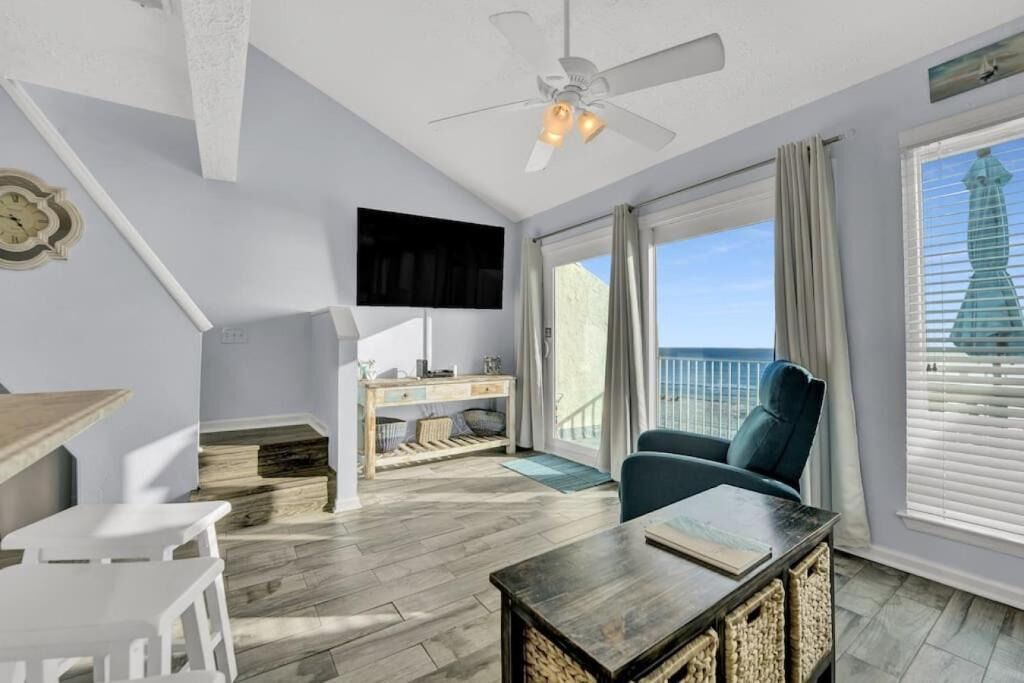 Quarto com ventilador de teto, televisão de tela plana, mesa de jantar com cadeiras, sofá e grande janela com vista para o oceano.