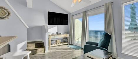 Quarto com ventilador de teto, televisão de tela plana, mesa de jantar com cadeiras, sofá e grande janela com vista para o oceano.