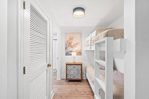 A narrow hallway with a wooden floor, white walls, a bunk bed, a decorative nightstand, and a ceiling light fixture. - Laketown Wharf (Panama City Beach)