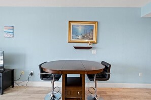 A dining area with a round table, two chairs, a painting on the wall, and a small shelf below the painting. - Laketown Wharf (Panama City Beach)